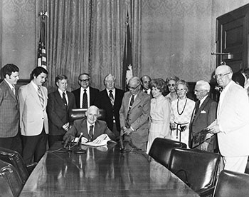 black and white photo of Governor John C. West signing the bill to make Winthrop coeducational.
                              He is seated at a long table and surrounded by a group of men and women