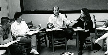 a black and white photo of Roger Baumgarte teaching a class. He is seated at a desk
                              in a circle with the students