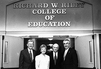 black and white photo of Richard W. Riley, Anthony DiGiorgio and others in front of
                              a sign with the new name for the college of education