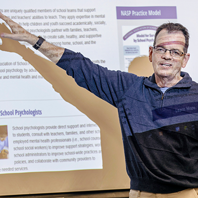 Caucasian male teacher pointing at a projection of a webpage that is describing school
                           psychologists