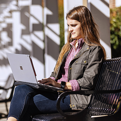 Woman sitting on a bench outside working on a laptop in her lap