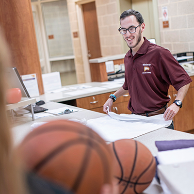 Man standing at a reception desk folding a towel while someone puts basketballs on
                           the table in the foreground
