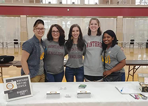 Five tutors stand smiling behind a table with Winthrop shirts